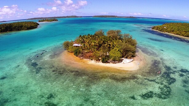 Eine Vogelperspektive einer tropischen Insel mit klarem türkisblauem Wasser, umgeben von üppiger Vegetation und einigen kleinen Gebäuden.