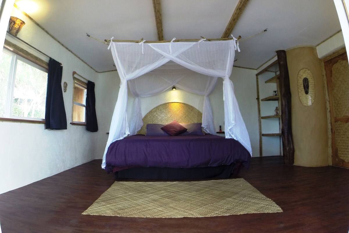 A room with a bed under a white canopy, a window with curtains, and a wooden floor.