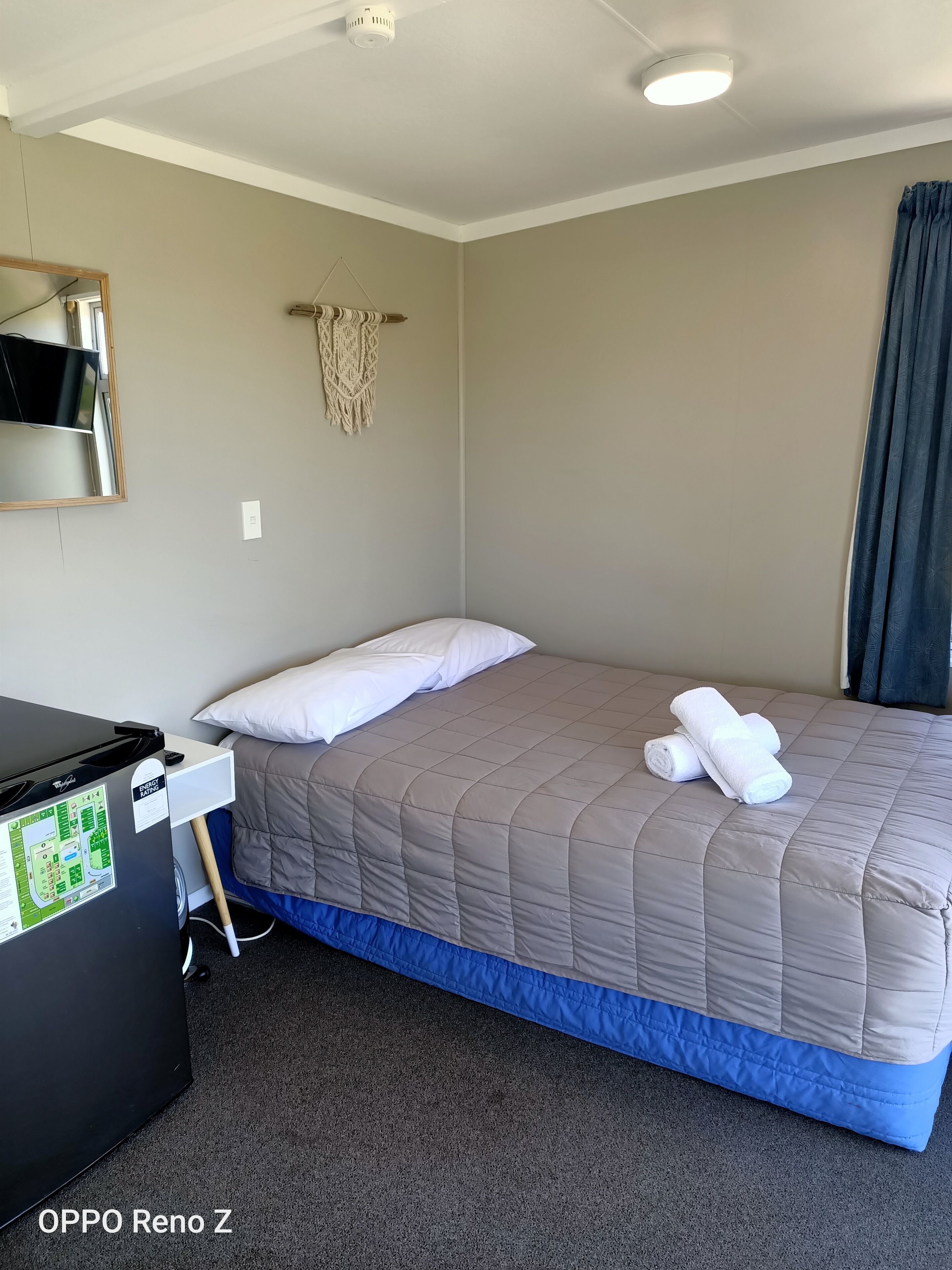 A neatly made bed with a beige quilt, white pillows, and rolled white towels. There is a small desk with a black appliance, possibly a coffee maker, and a map on it. The room has a simple, clean design with a light-colored wall and a dark curtain on one side.