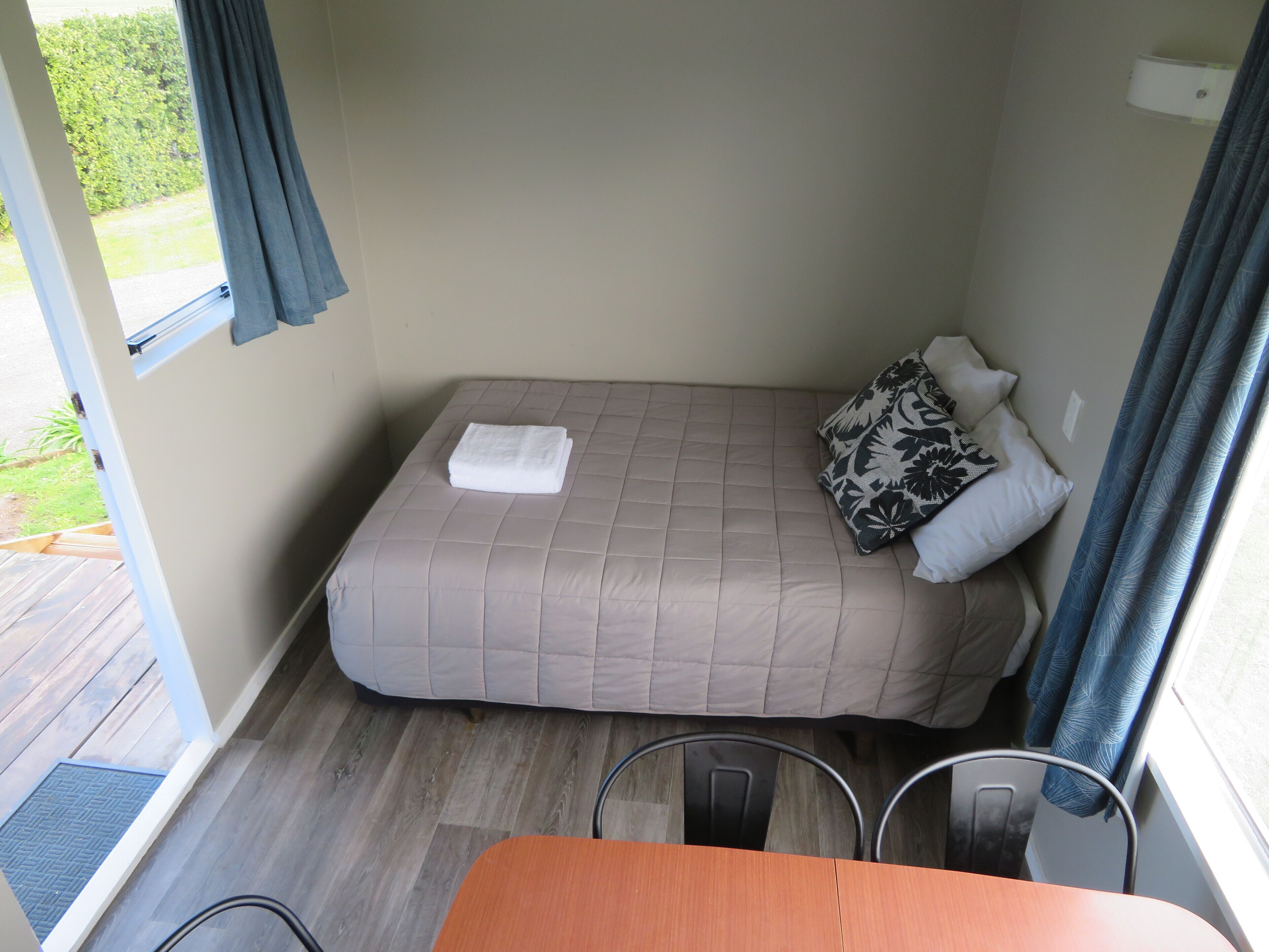 Ein kleines Zimmer mit einem Einzelbett, Holzboden, einem Tisch und einem Fenster mit Vorhängen.