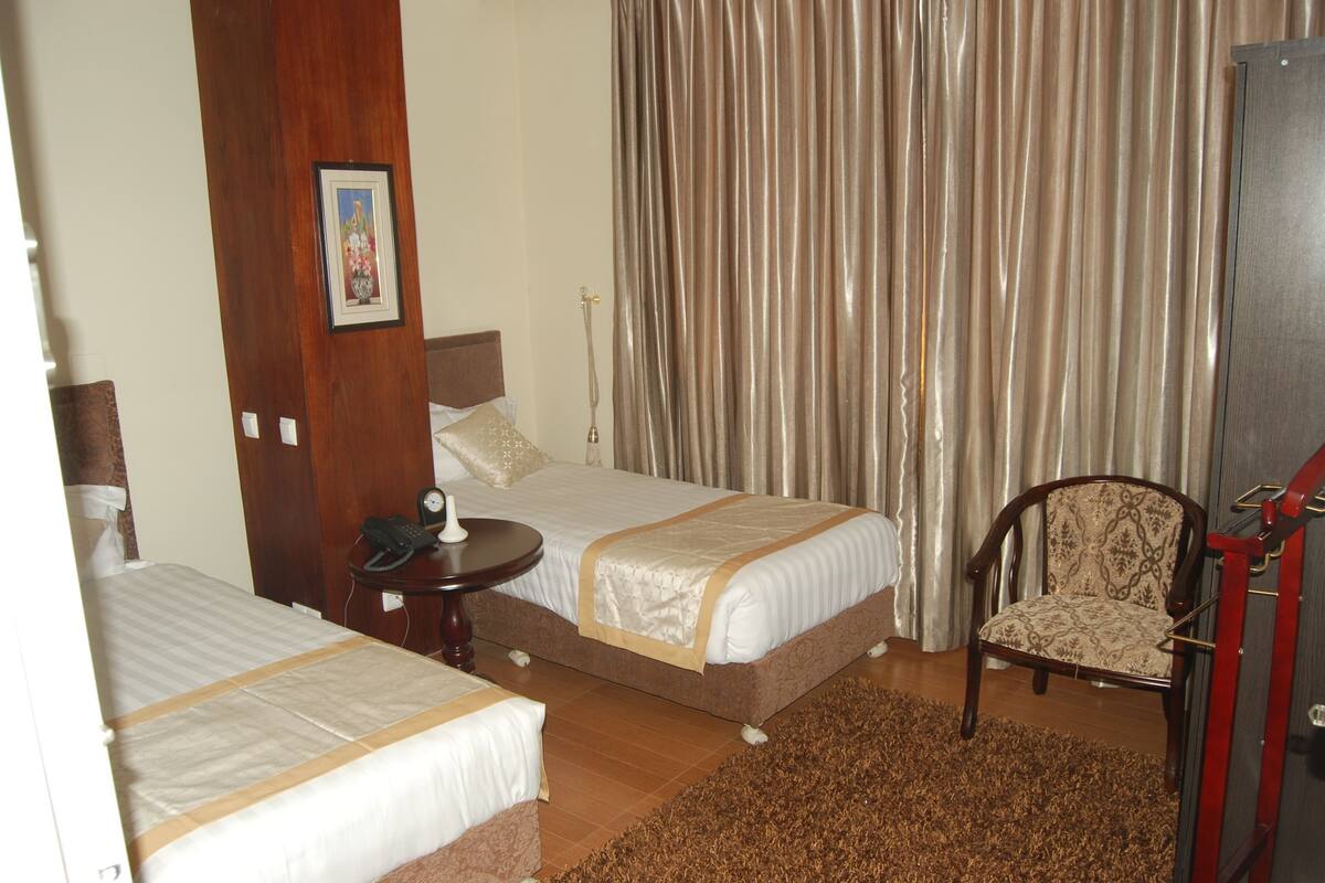 A hotel room with two beds, a chair, a small table, and a curtain.