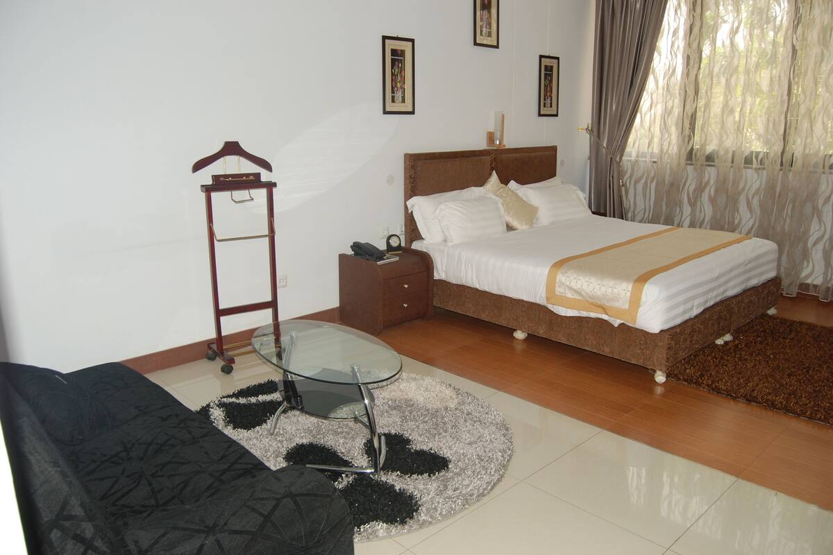 A bedroom with a bed, a glass table, a hanging coat rack, and a black sofa.