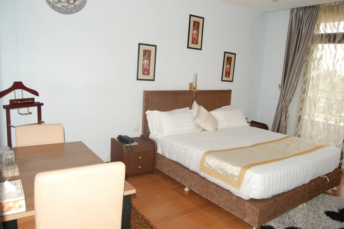 A hotel room with a large bed, a wooden nightstand, a desk with a chair, and a small table with glasses.