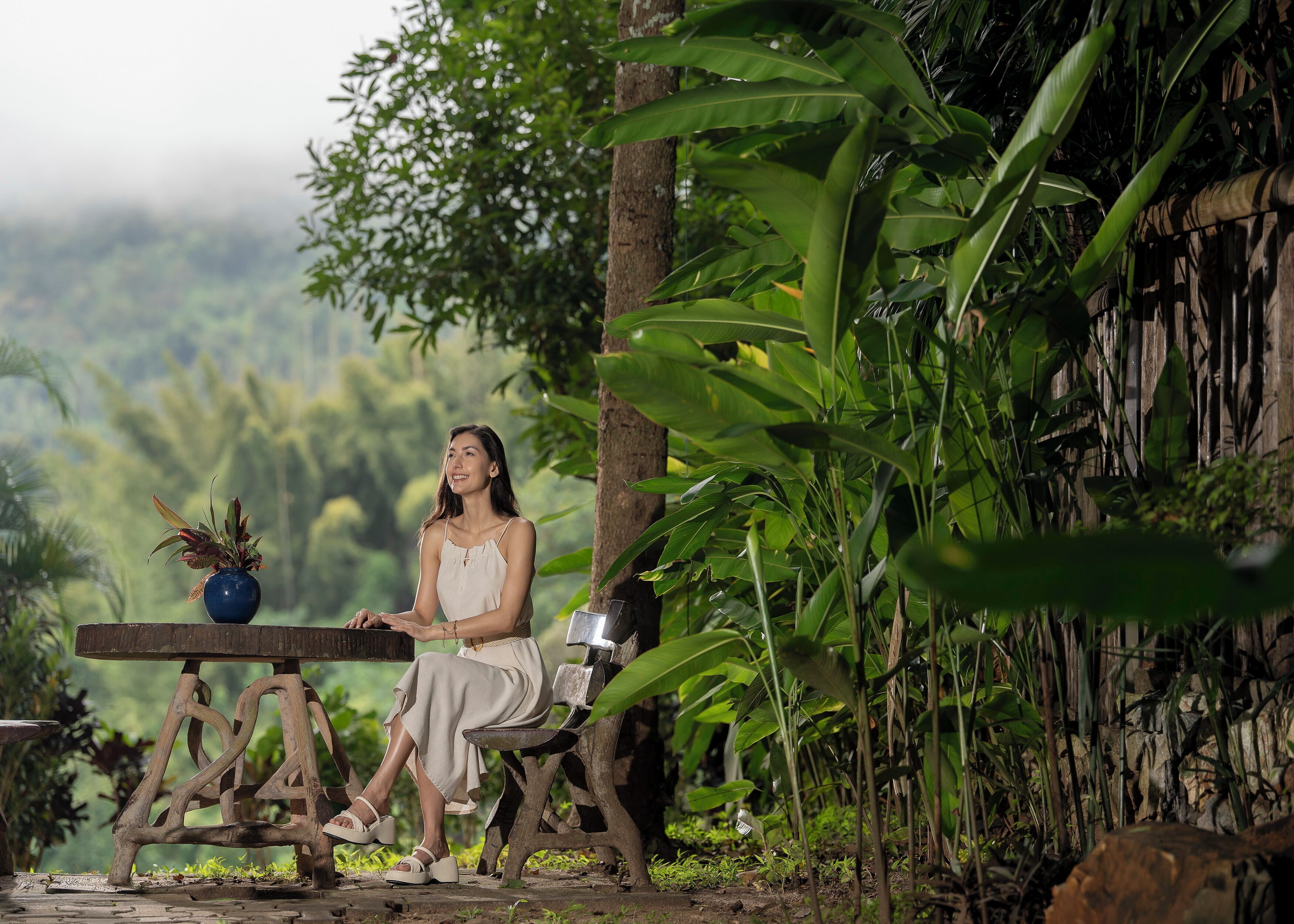 Une personne est assise sur un banc en bois à l’extérieur, avec un pot de fleurs à ses côtés, entourée d’une végétation luxuriante et d’une structure en bois.