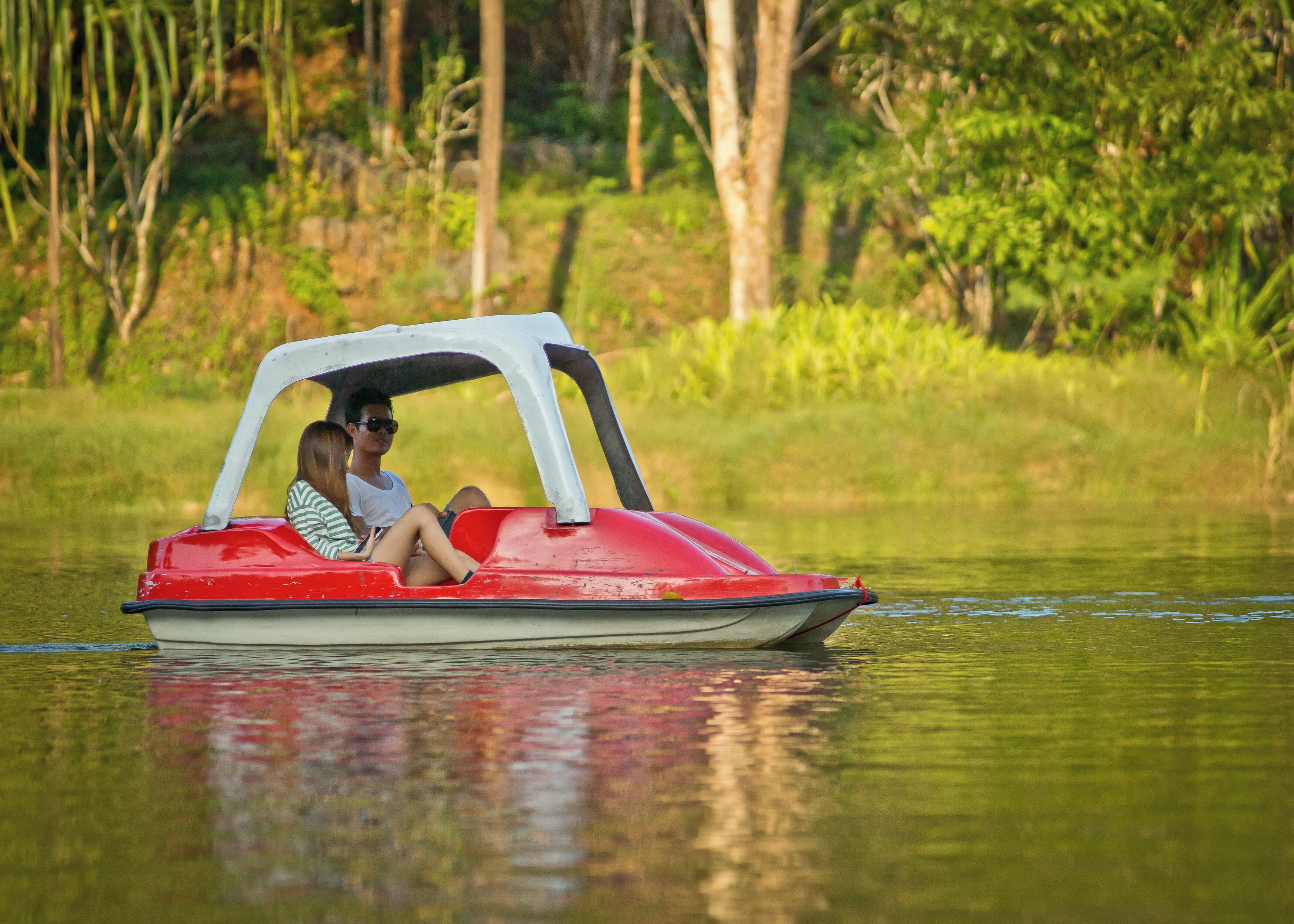 Un barco rojo y blanco con dos personas navegando sobre aguas tranquilas, rodeado de vegetación.