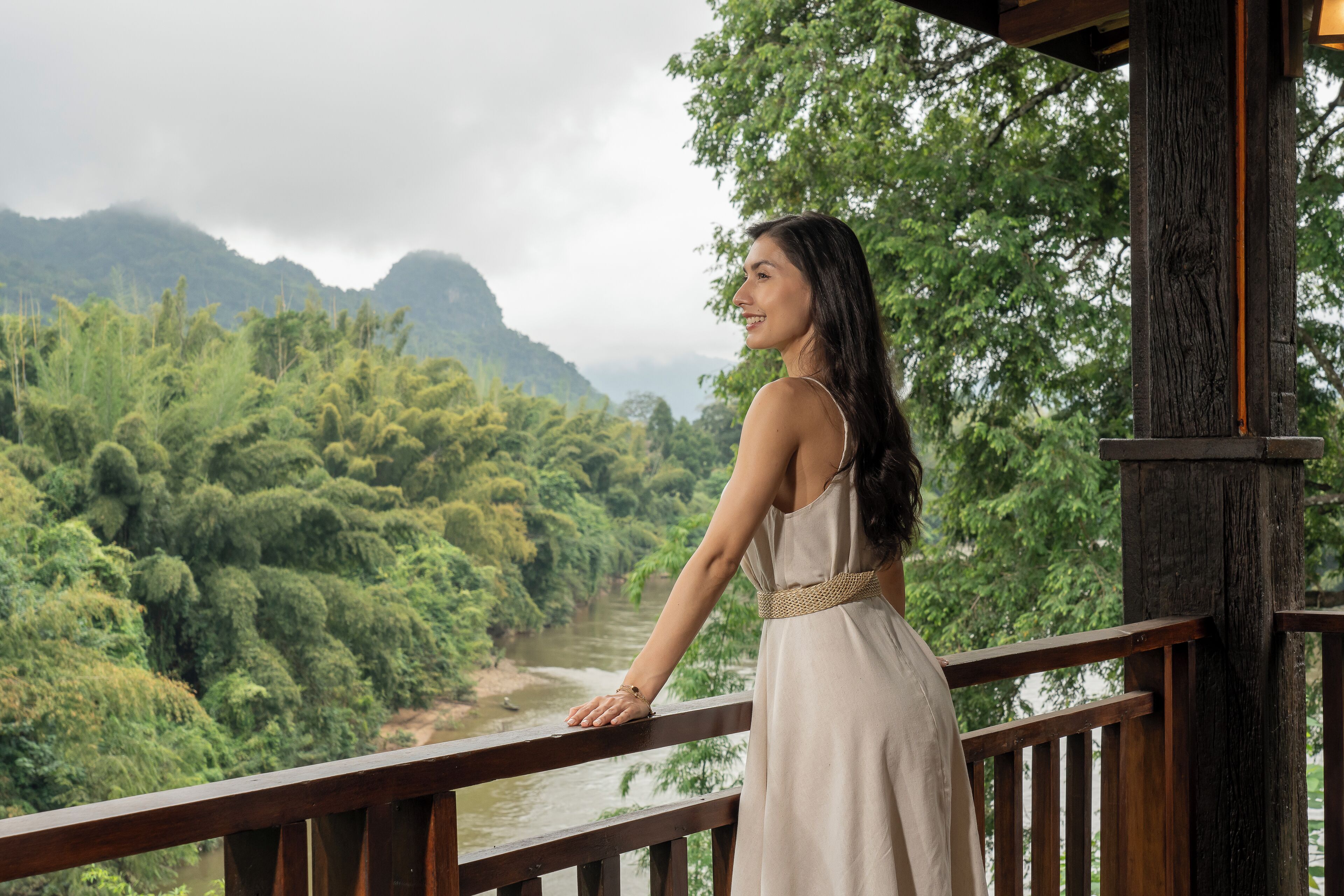 A woman in a dress standing on a wooden balcony overlooking a river and lush greenery.