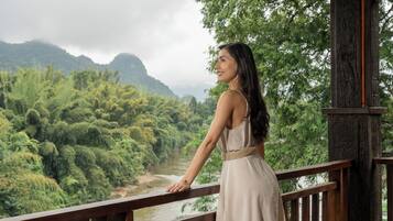 A woman in a dress standing on a wooden balcony overlooking a river and lush greenery.