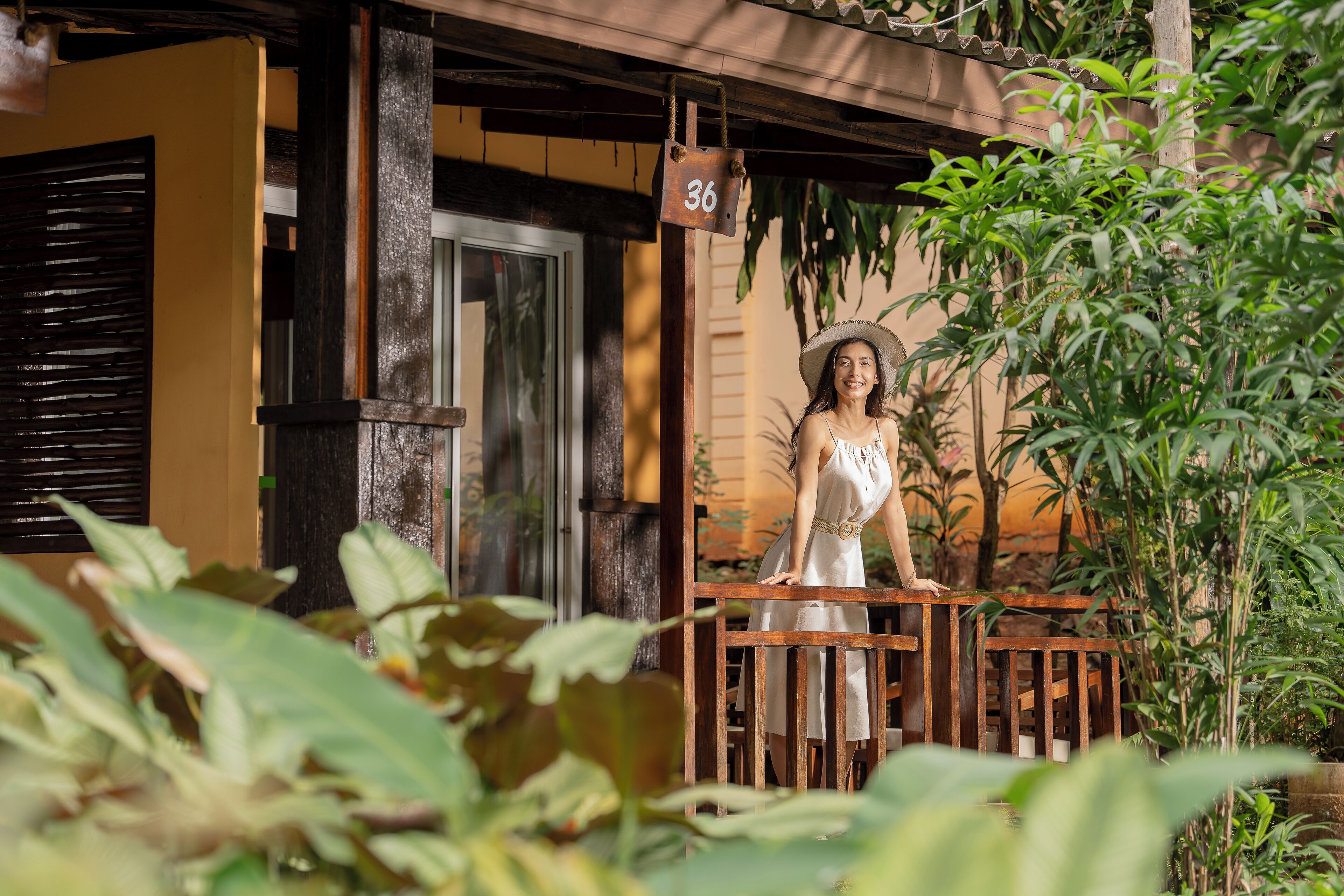 A woman standing on a wooden balcony with a number sign '36' above, surrounded by lush greenery.
