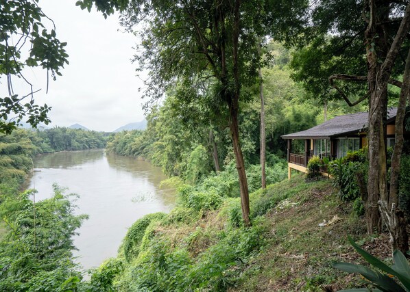 Vista al río con una pequeña casa de madera junto al agua, rodeada de abundante vegetación.