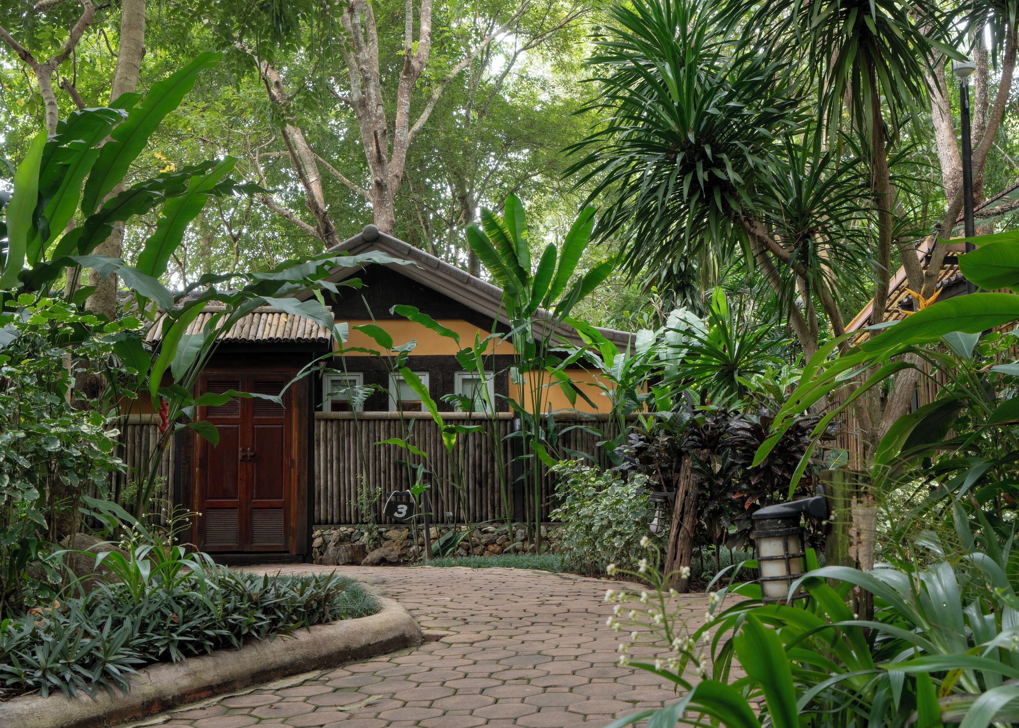 Une maison traditionnelle en bois, avec un toit de tuiles, entourée d’une végétation luxuriante et un chemin de pierres menant à l’entrée.