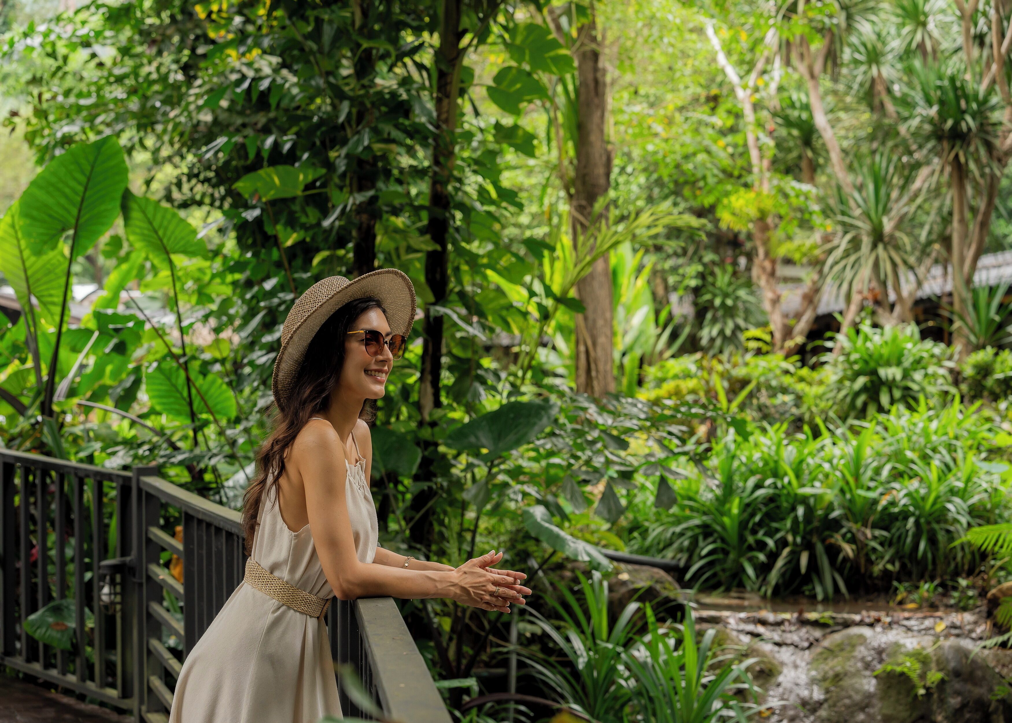 Una mujer con un vestido blanco y sombrero de paja, de pie junto a una baranda en un jardín exuberante, verde y lleno de follaje, con un pequeño arroyo.
