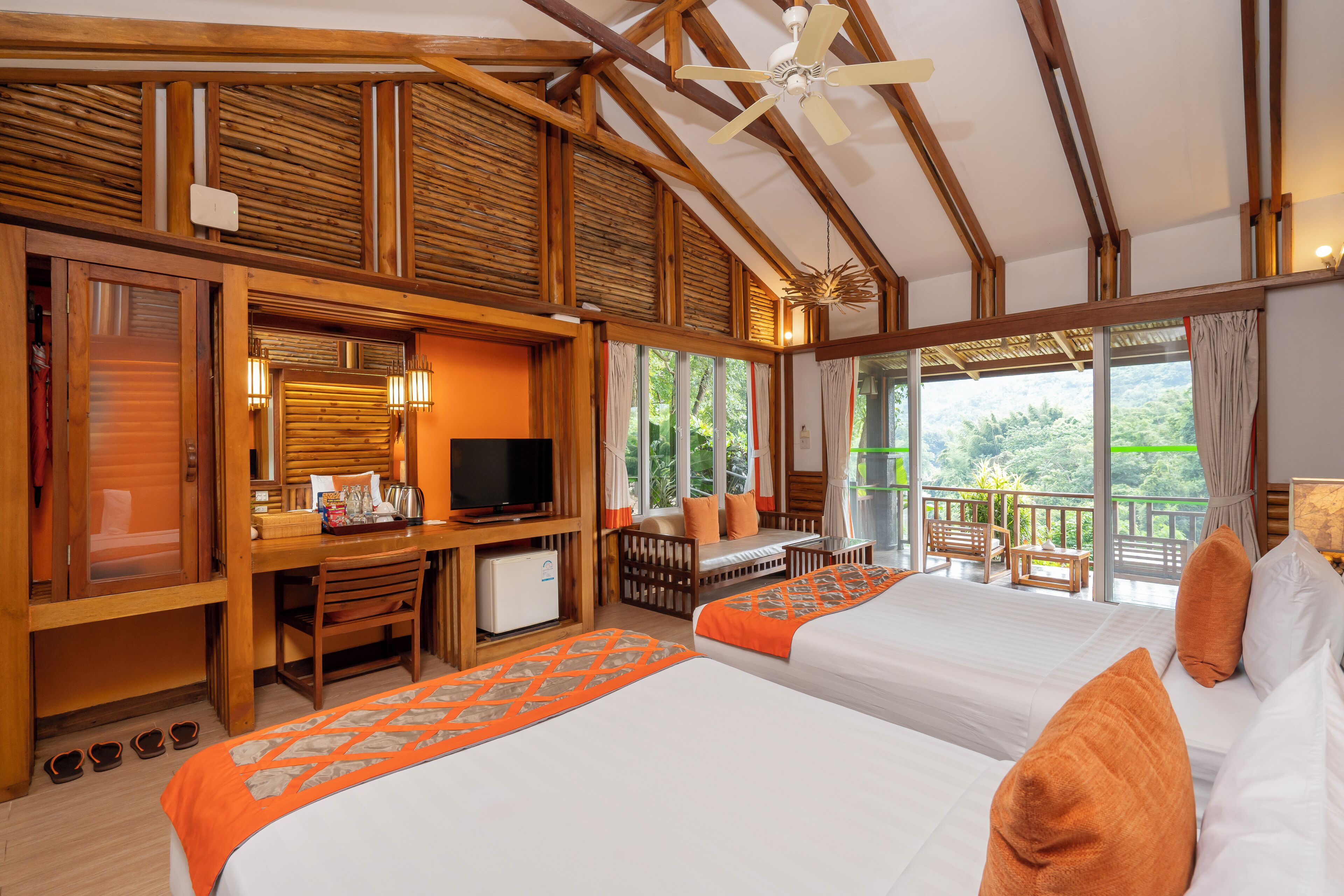 A hotel room with two beds, a desk with a television, a balcony with a view of the outdoors, and wooden architecture.