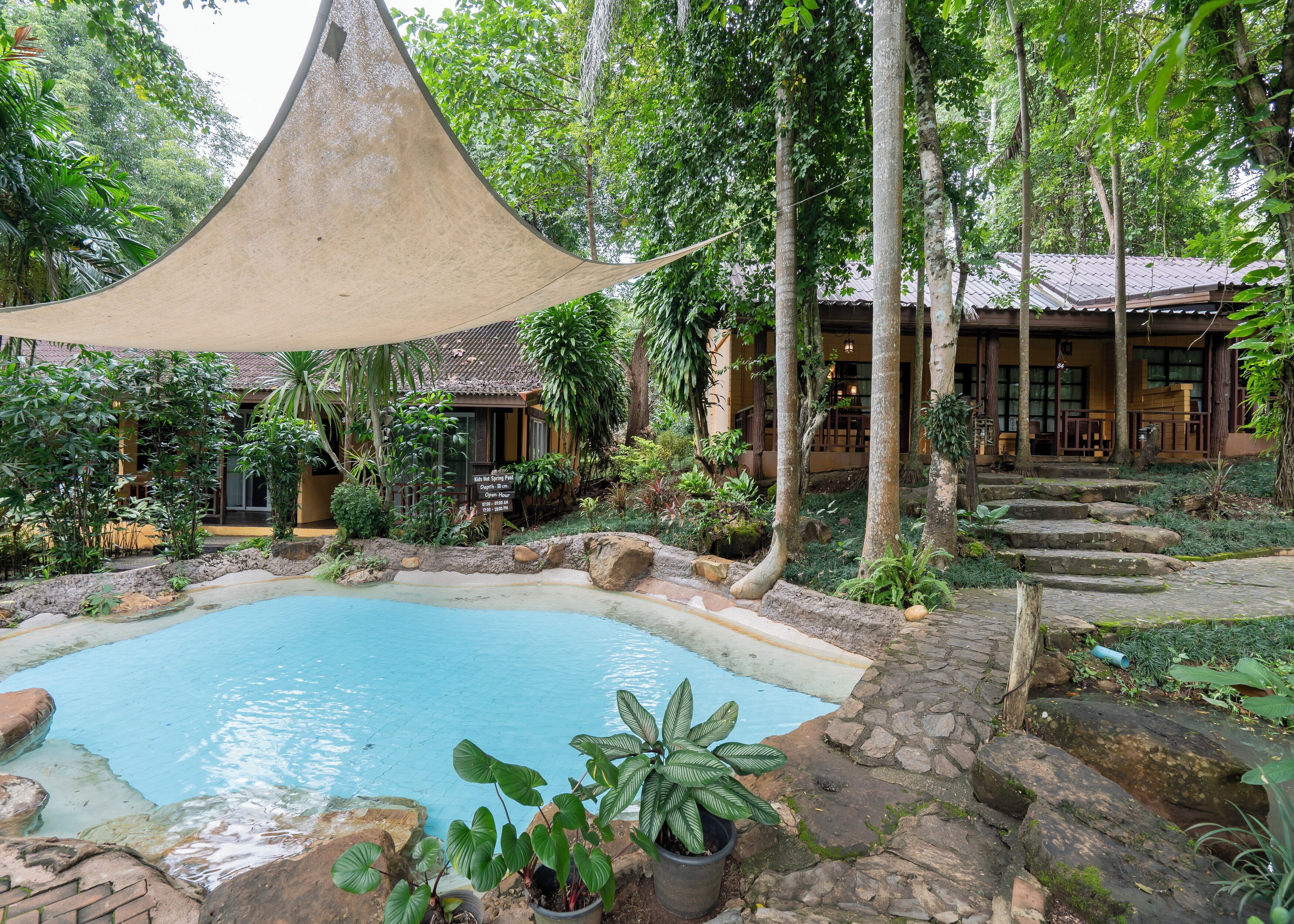 A pool area with a canopy, surrounded by tropical plants and stone steps leading to a building with a tiled roof.