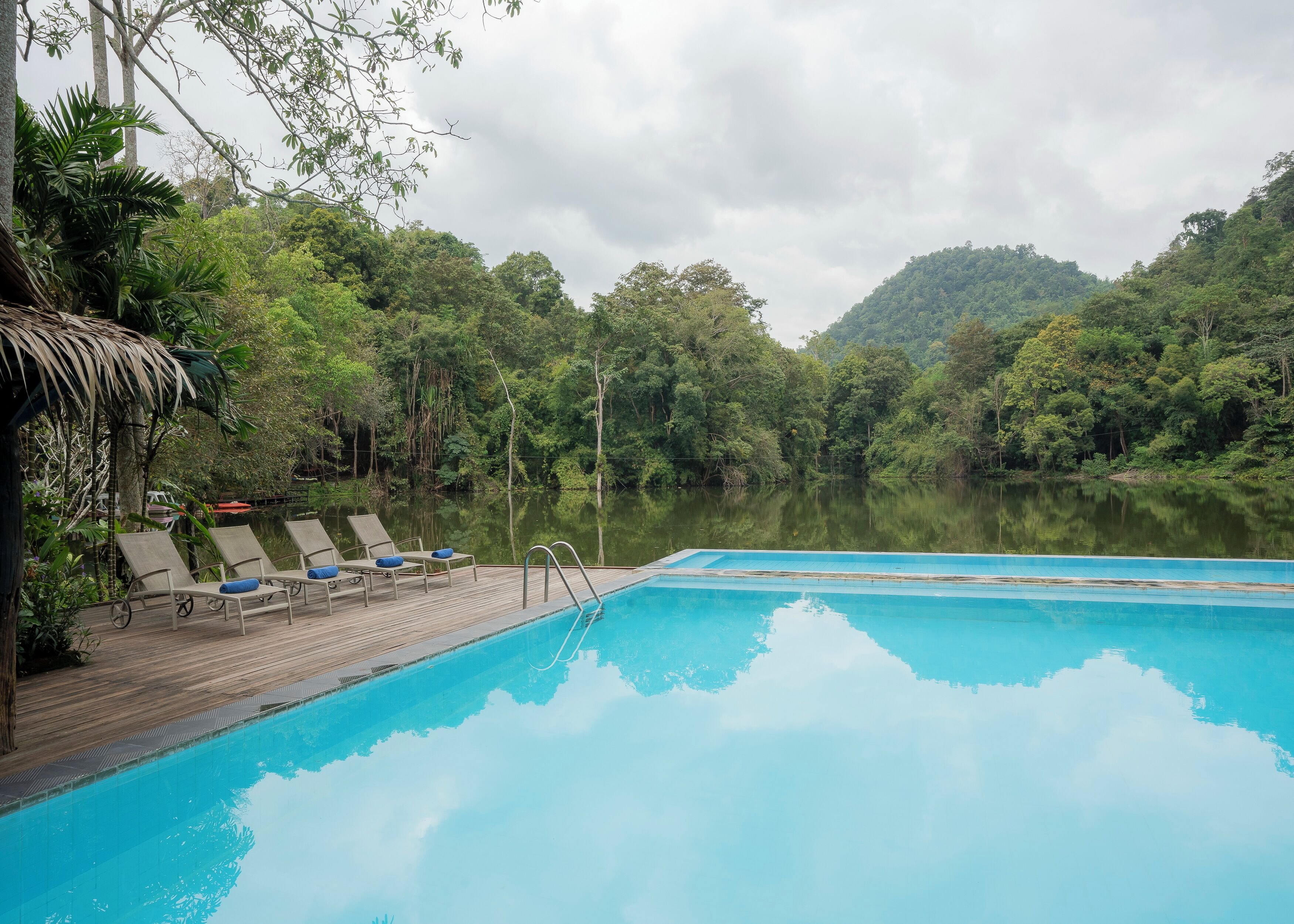Una piscina con lettini e un pontile in legno, immersa nel verde lussureggiante e affacciata su un lago tranquillo, con dolci colline sullo sfondo.