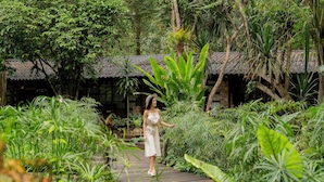 A woman walking on a wooden bridge surrounded by lush greenery and a traditional building with a tiled roof.