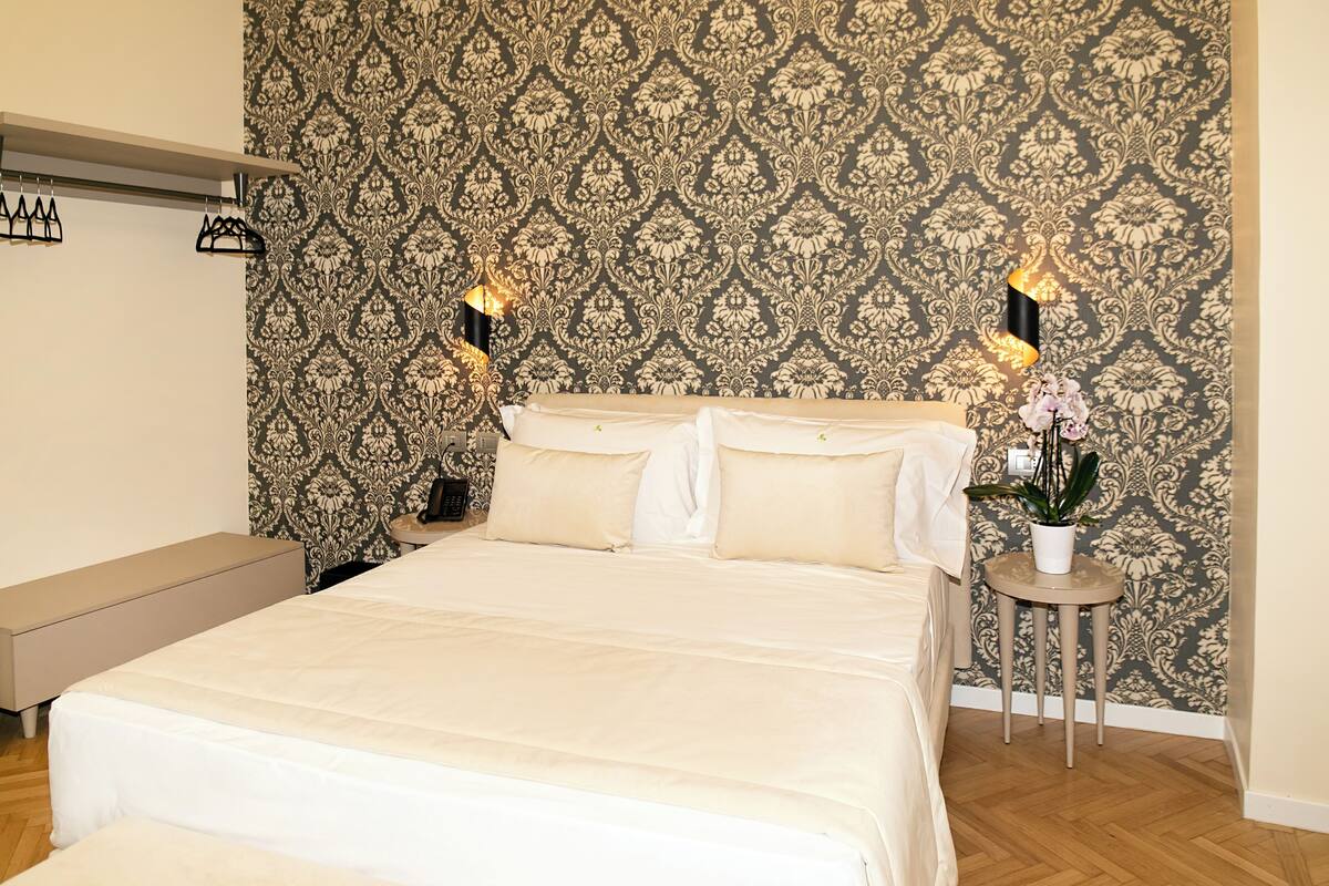 A neatly made bed with a white duvet, a bedside table with a lamp, and a small potted plant.