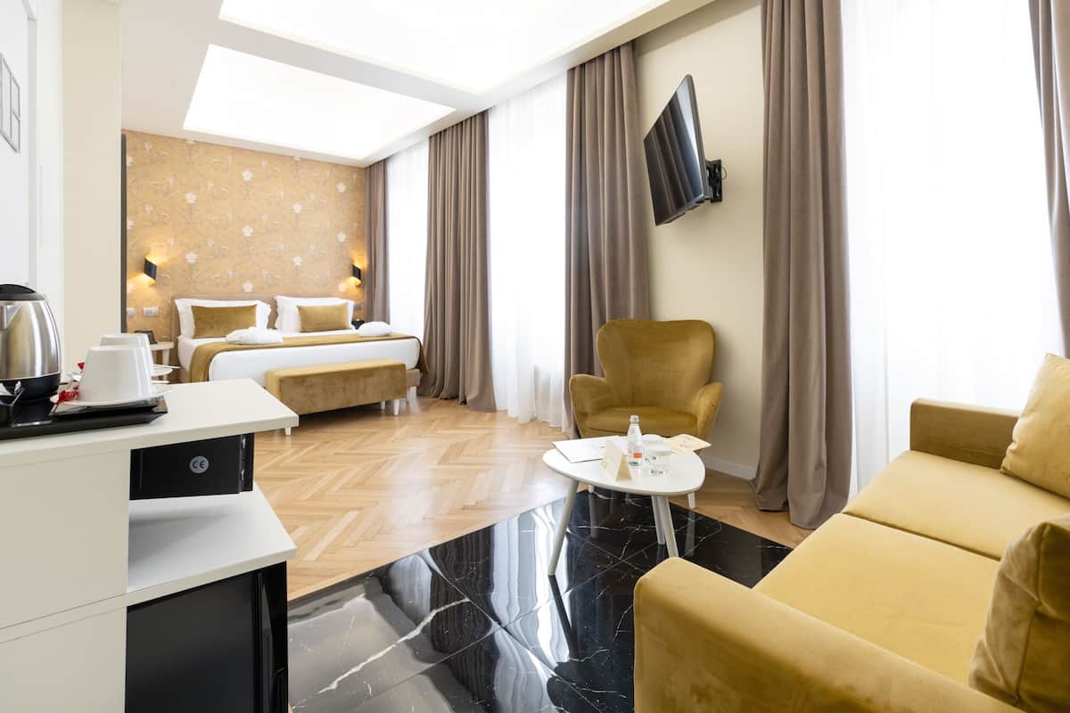 A hotel room with a bed, a sofa, a chair, a coffee table, a TV, and a desk with a kettle and cups.