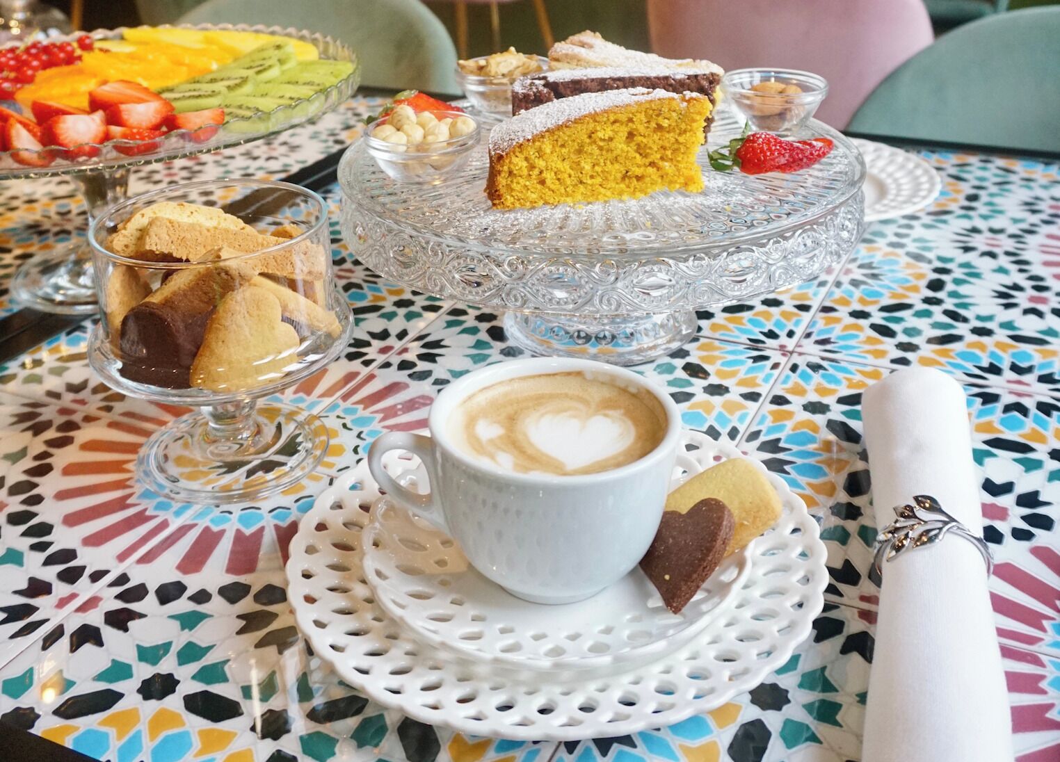 A table with a colorful mosaic tablecloth, a glass cake stand with a slice of yellow cake, a cup of coffee with latte art, and a white napkin holder.