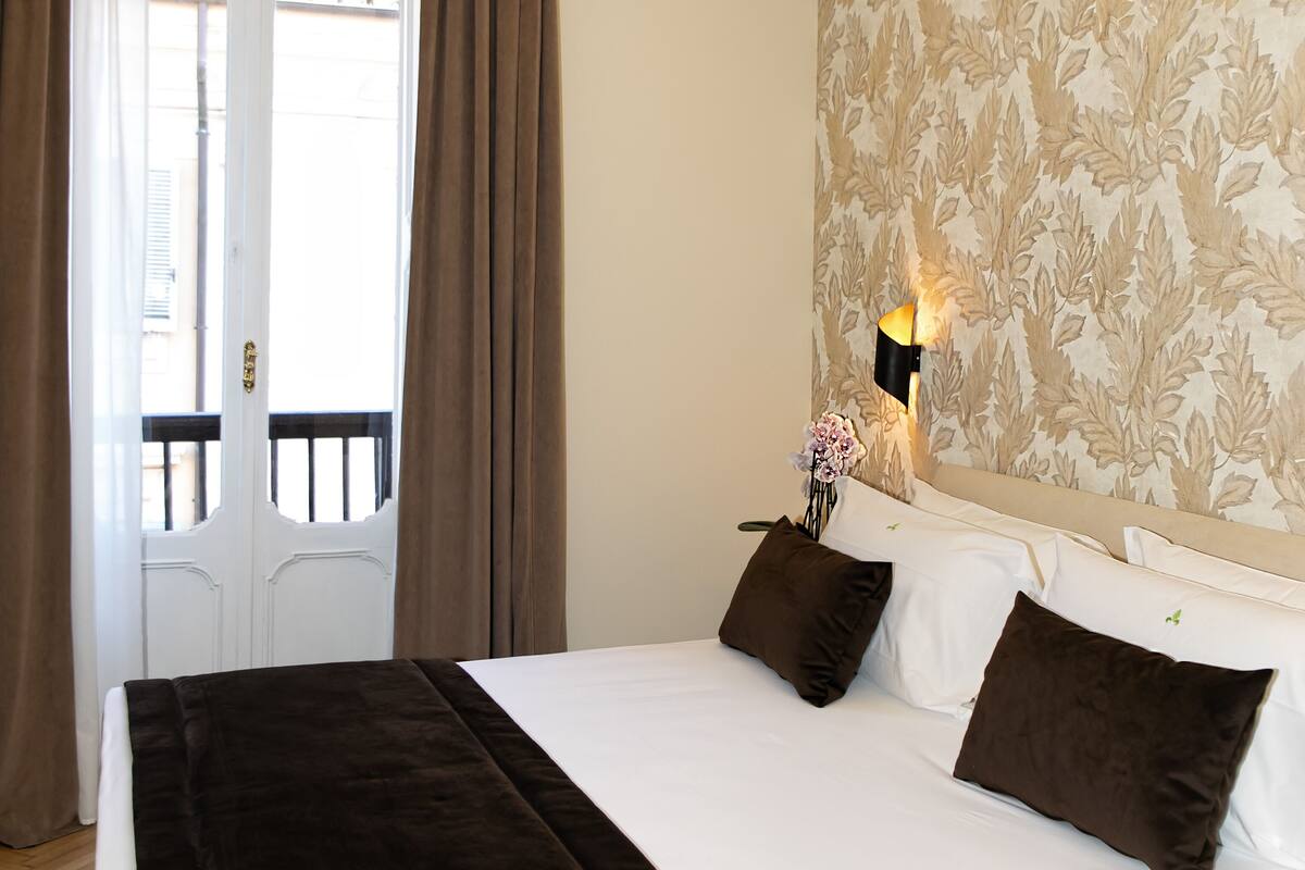 A hotel room with a large bed, a nightstand with a lamp, a window with curtains, and a patterned wall.