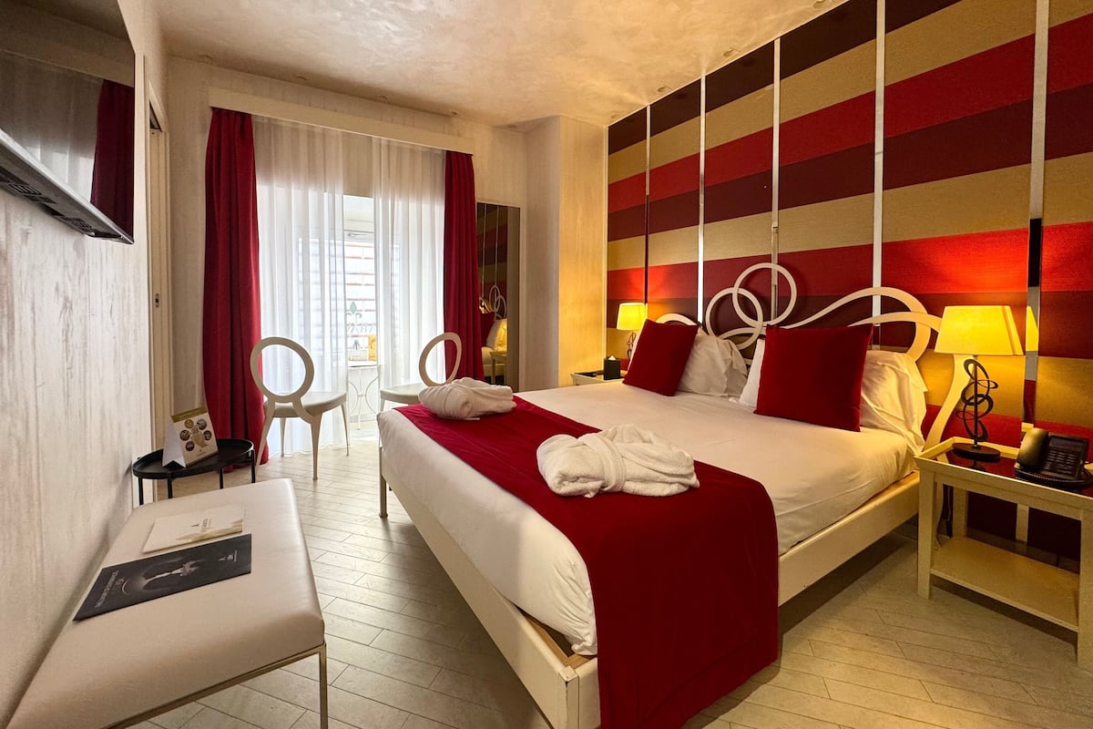 A hotel room with a large bed, two bedside tables with lamps, a desk with a chair, and a window with curtains.