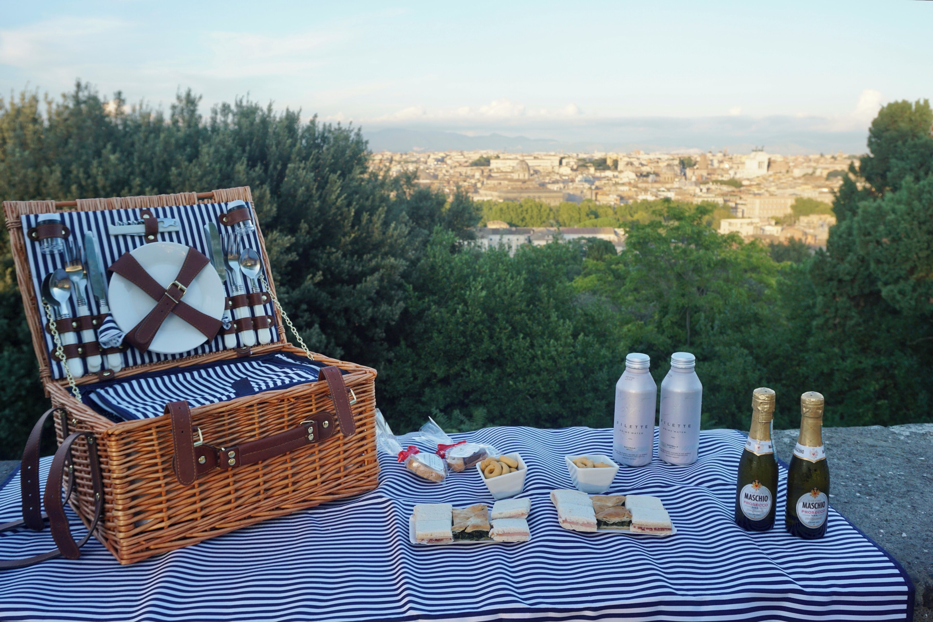 A picnic setup with a wicker basket, sandwiches, pastries, and bottles of champagne on a striped blanket with a city view in the background.