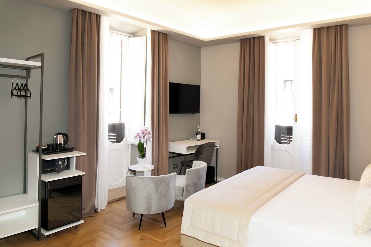 A hotel room with a bed, desk, chair, TV, and a small table with a flower vase.