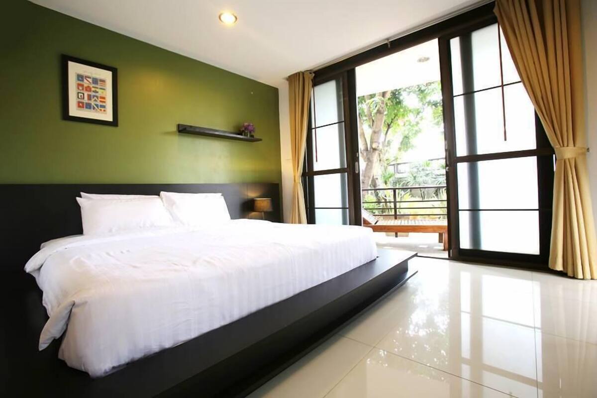 Een slaapkamer met een groot bed, een groene wand, een raam met gordijnen en een klein plankje met een plant.