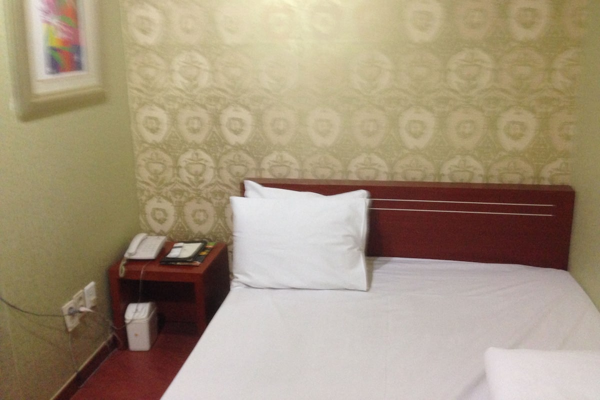 A hotel room with a patterned wallpaper, a wooden headboard, a white pillow, a bedside table with a phone, and a small trash bin.
