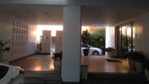 Entrada de un edificio moderno con un porche cubierto, plantas en macetas y un coche blanco estacionado afuera.