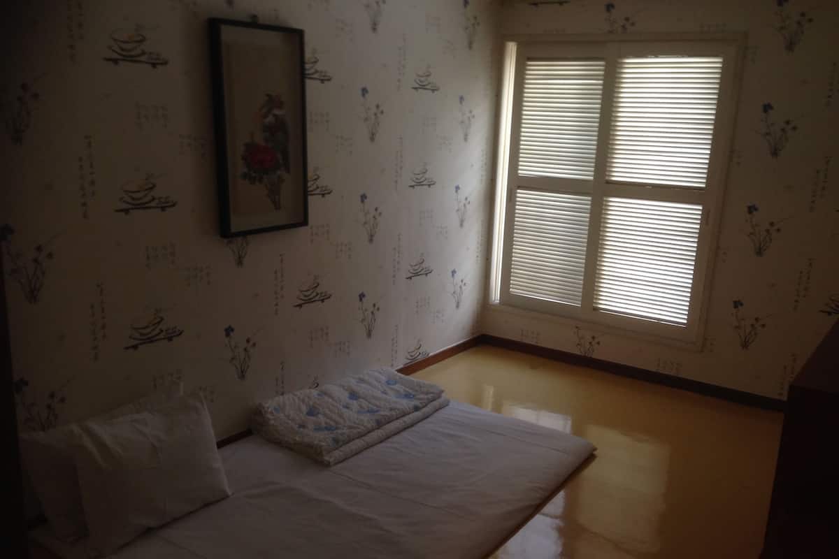 A room with a bed, a window with blinds, and a wall with floral patterns.