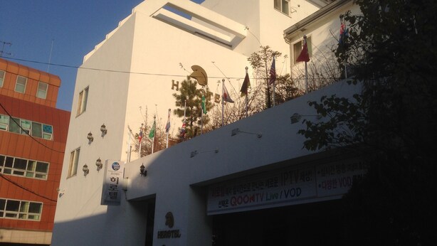 Un hotel con fachada blanca, varias banderas y un letrero para QOOMTV Live/VOD.