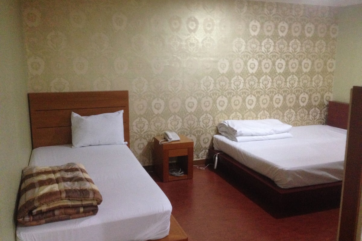 A hotel room with two beds, a wooden headboard, a bedside table with a phone, and patterned wallpaper.