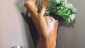 Un rincón con una escultura de madera, una planta en maceta y un cubo de basura.