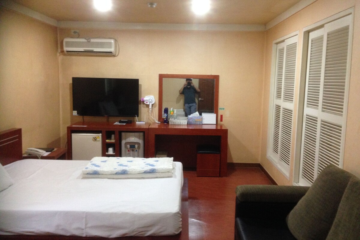 Una habitación de hotel con cama, escritorio con televisor de pantalla plana, espejo, dispensador de agua y armario con puertas batientes.