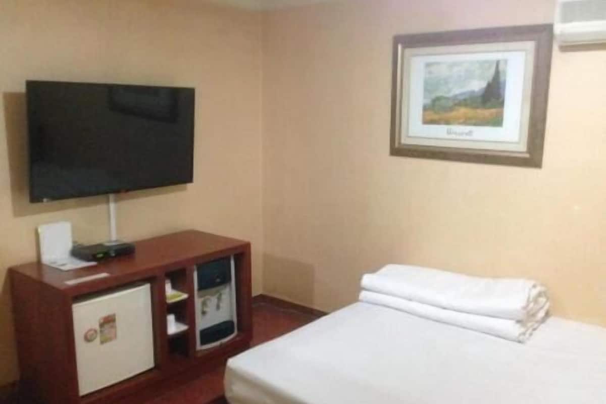 A hotel room with a bed, a TV mounted on the wall, a small cabinet with a refrigerator, and a framed picture on the wall.