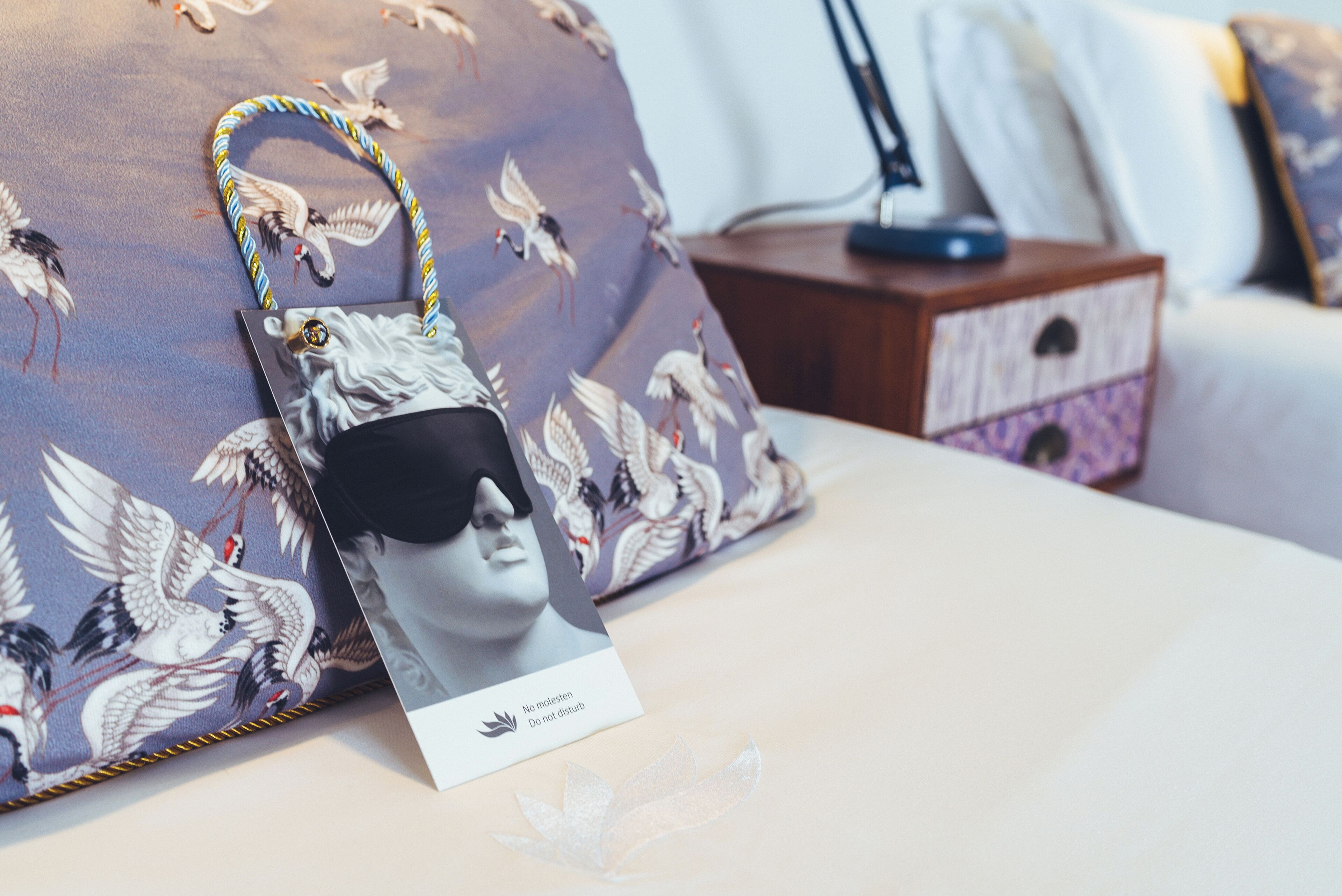 Una cama con un cojín estampado con un diseño de pájaro, una mesita de noche con una lámpara y una tarjeta con un mensaje.