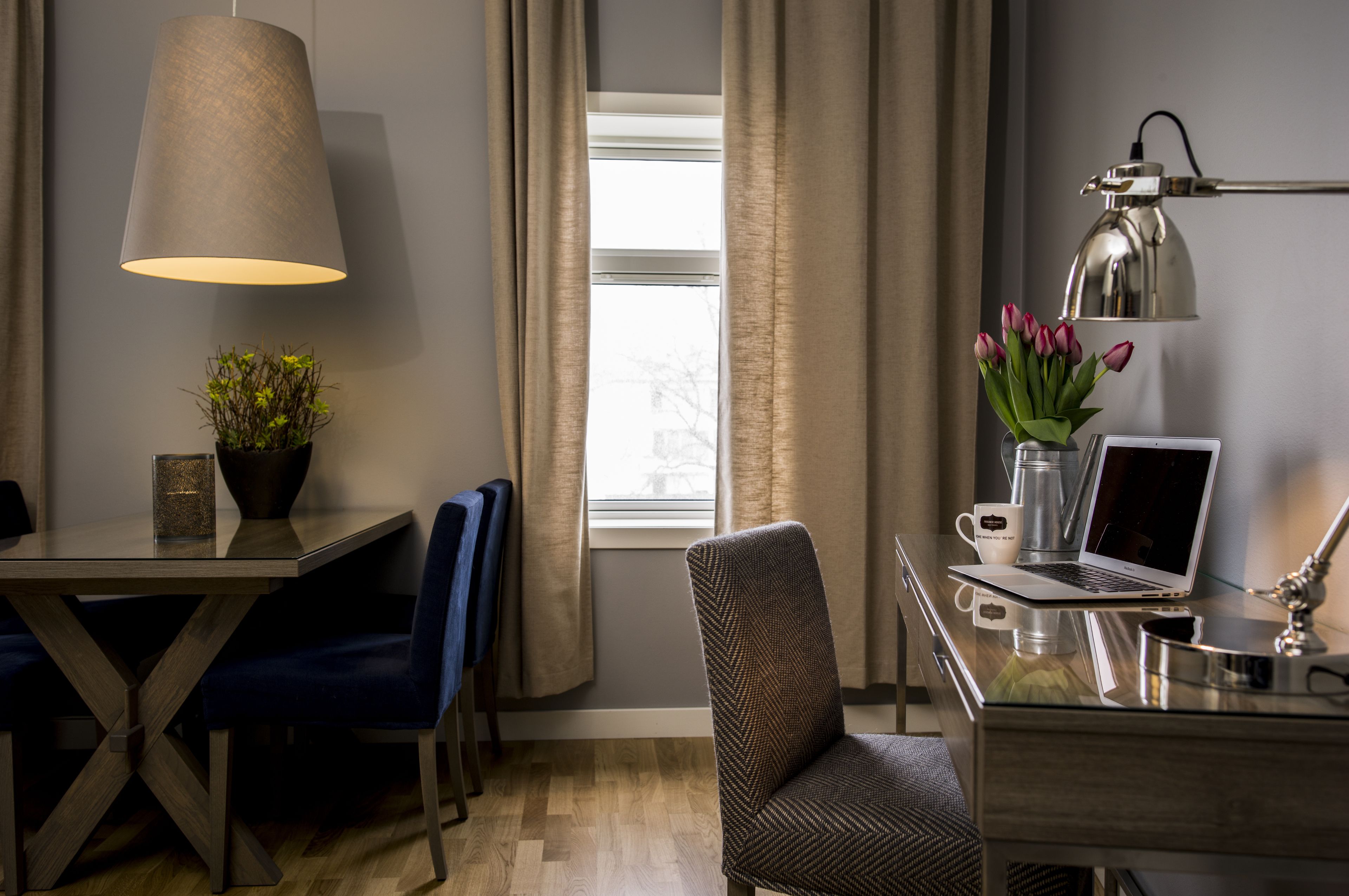 Una habitación de hotel moderna con una mesa de comedor de madera, sillas azules, una computadora portátil sobre un escritorio, una lámpara, una cafetera y un florero con flores.