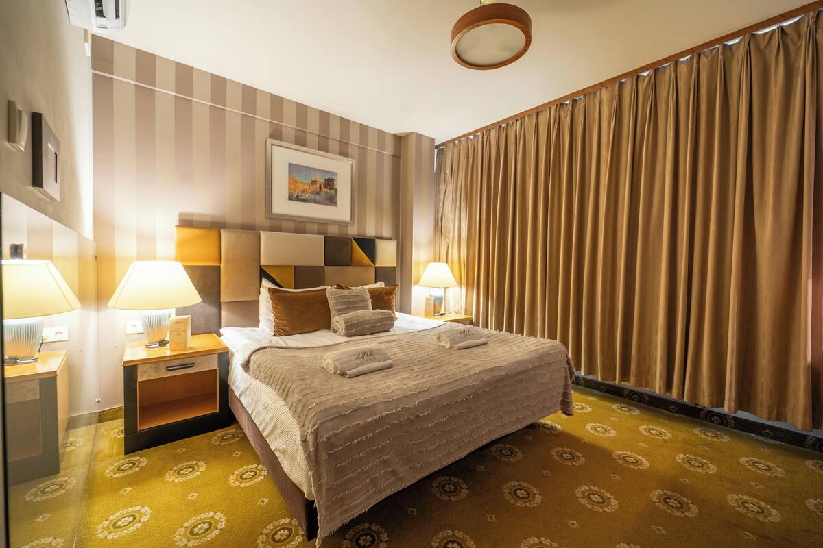 A hotel room with a large bed, two bedside tables with lamps, a headboard with a geometric pattern, a framed picture on the wall, and a large window with curtains.
