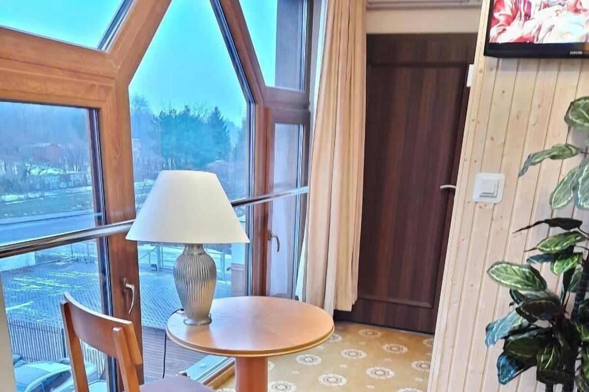 A room with a large window, a table with a lamp, a chair, a potted plant, and a bed.