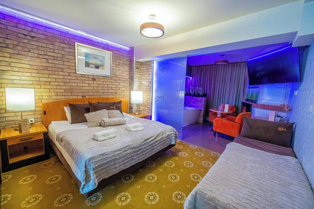 A hotel room with a brick wall, a bed with a white comforter and pillows, a small table with a lamp, and a TV mounted on the wall.