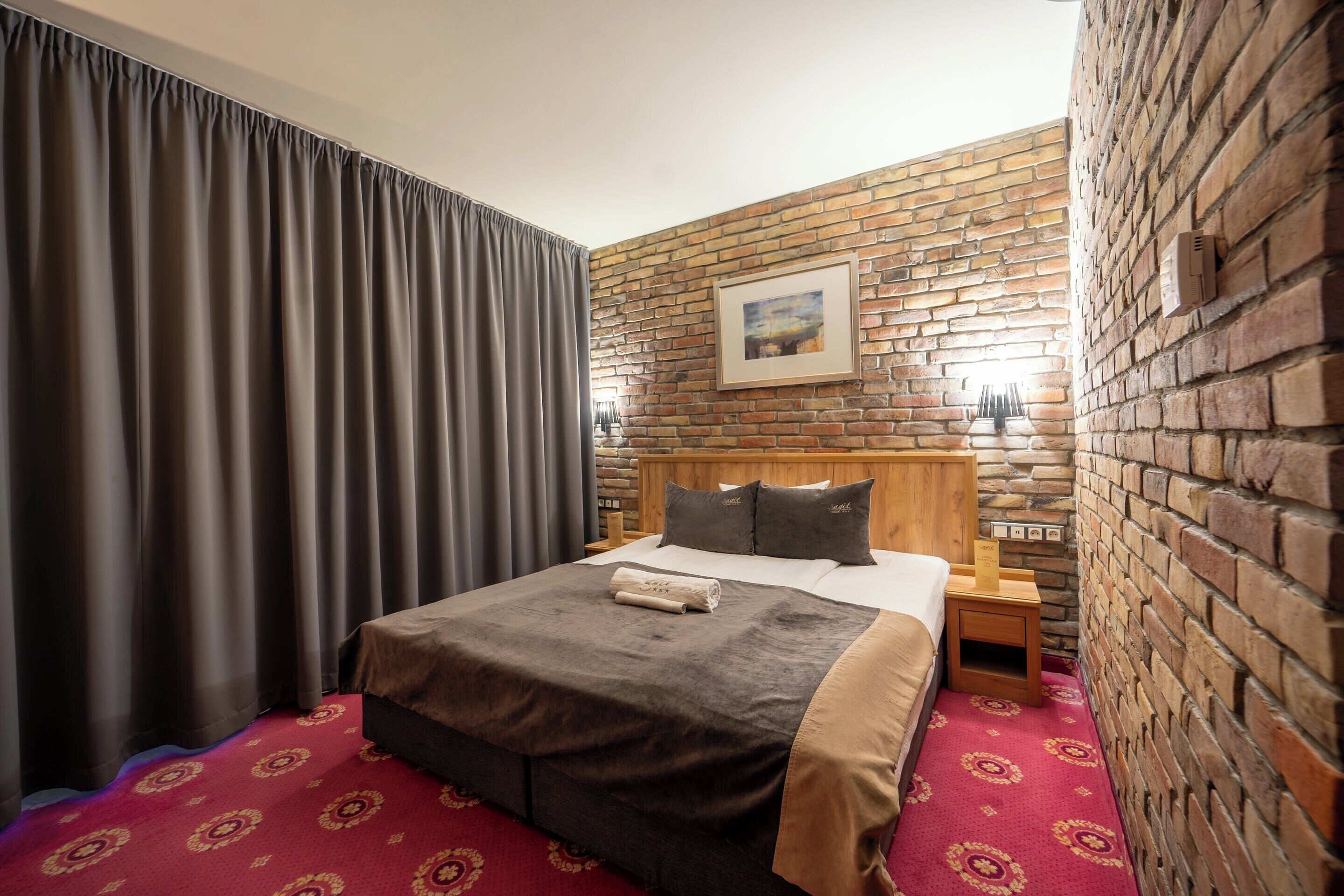 Habitación de hotel con pared de ladrillo, cama grande con cabecera de madera, mesita de noche y alfombra roja con estampado.