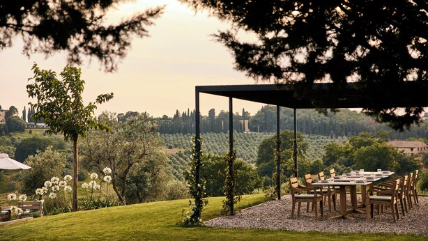 Un espacio exterior para comer con mesa y sillas, rodeado de follaje y árboles, con vistas a un paisaje pintoresco.