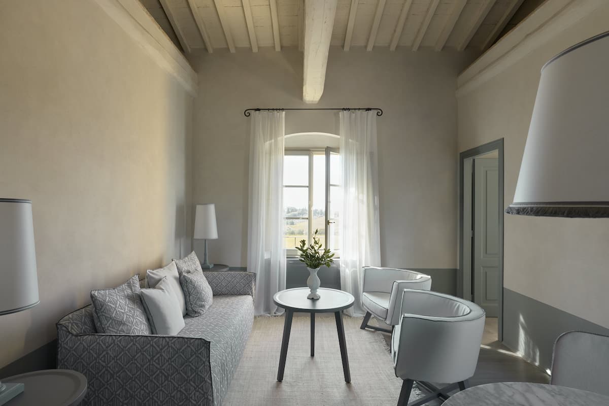 A modern hotel room with a sofa, two armchairs, a small table, and a view through a large window.