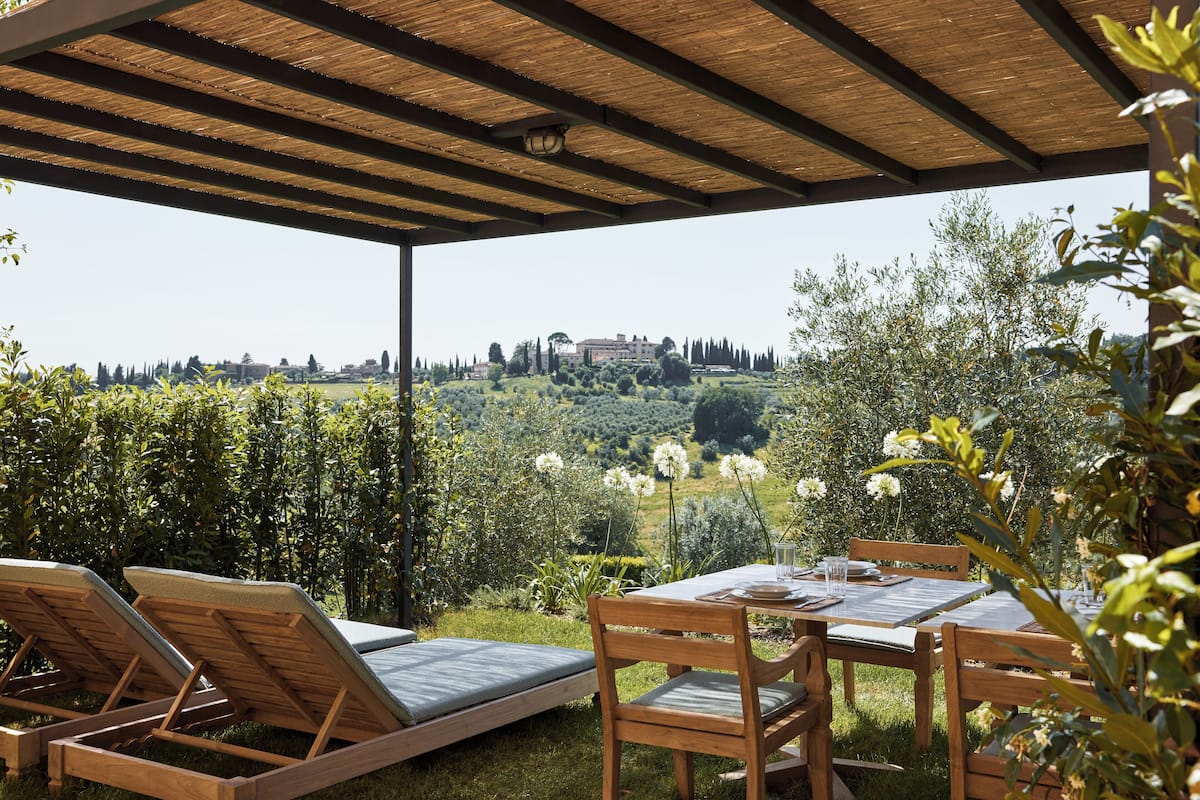 A covered outdoor seating area with wooden furniture and a table, overlooking a lush landscape with trees and a distant building.