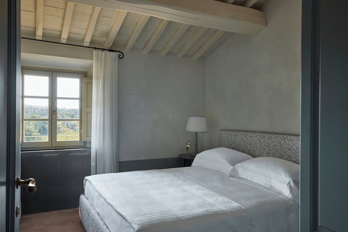A bedroom with a bed, a nightstand with a lamp, a window with curtains, and a view of greenery outside.