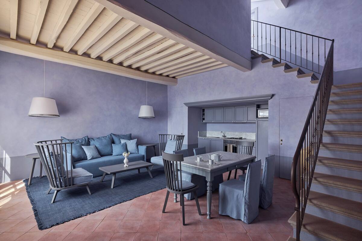 A modern living room with a sofa, chairs, a dining table, and a kitchen area with cabinets and a staircase.