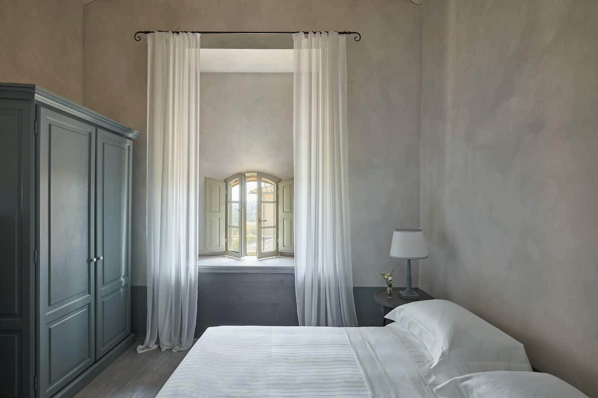 A bedroom with a large bed, a wardrobe, a nightstand with a lamp, and a view of a window and a door.