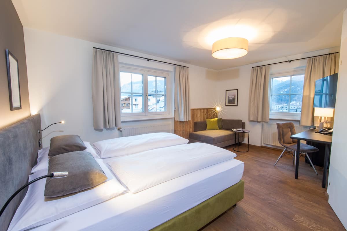 A hotel room with a double bed, a desk with a chair, a sofa, and a view of buildings through the windows.