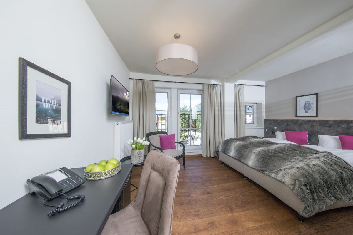 A hotel room with a bed, a desk with a phone and a bowl of apples, a chair, and a large window with curtains.