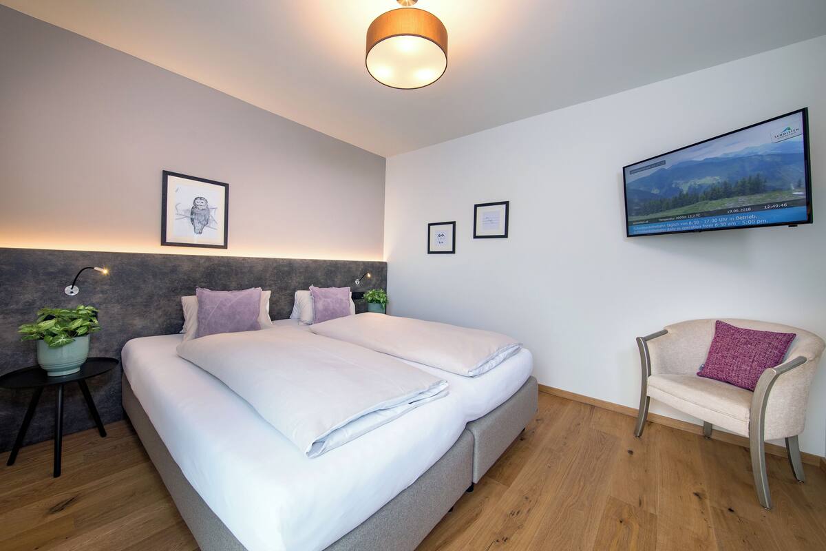 A modern hotel room with a large bed, a small side table with a plant, a sofa, and a wall-mounted TV.